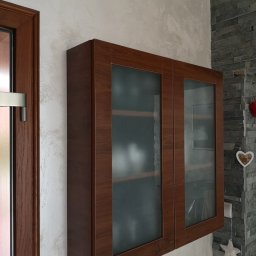 KEM Marcin Wielicki - Wisząca szafka z brązowymi ramami i matowymi szklanymi drzwiczkami, zamontowana na ścianie z dekoracyjną fakturą, obok ściany z szarej okładziny kamiennej i ozdobami w kształcie serca i gwiazdy.