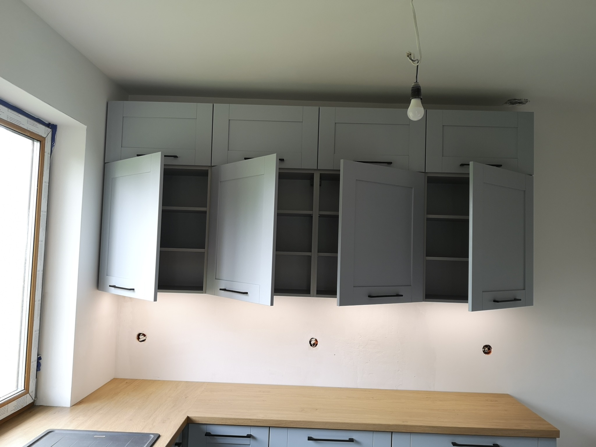 Szare szafki kuchenne z drewnianym blatem, niektóre fronty otwarte, widoczna instalacja elektryczna na ścianie, surowa żarówka zwisająca z sufitu.