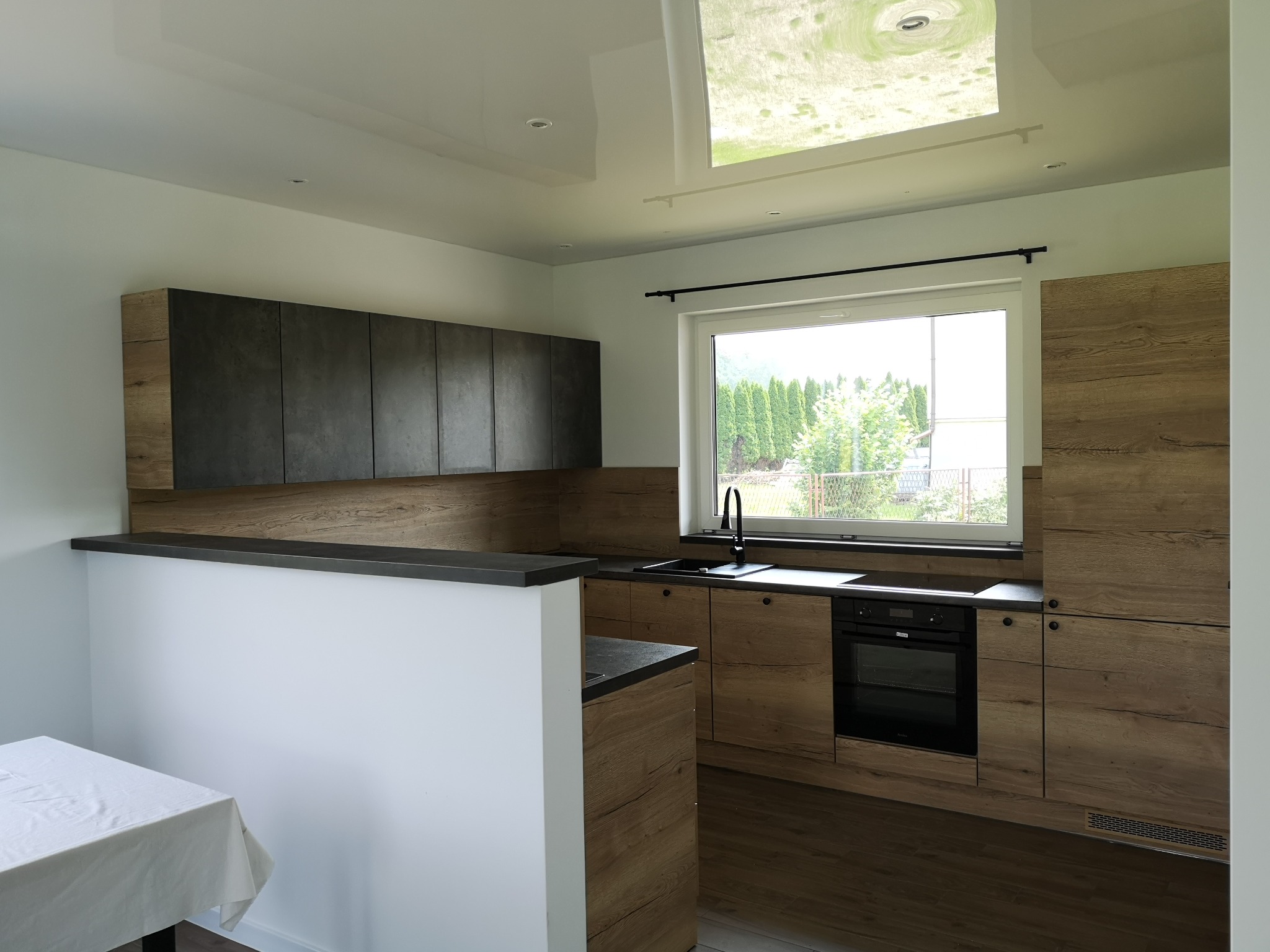 Jasne wnętrze kuchni z drewnianymi szafkami dolnymi i ciemnoszarymi górnymi, widok na ogród przez okno, biały stół z lewej strony.