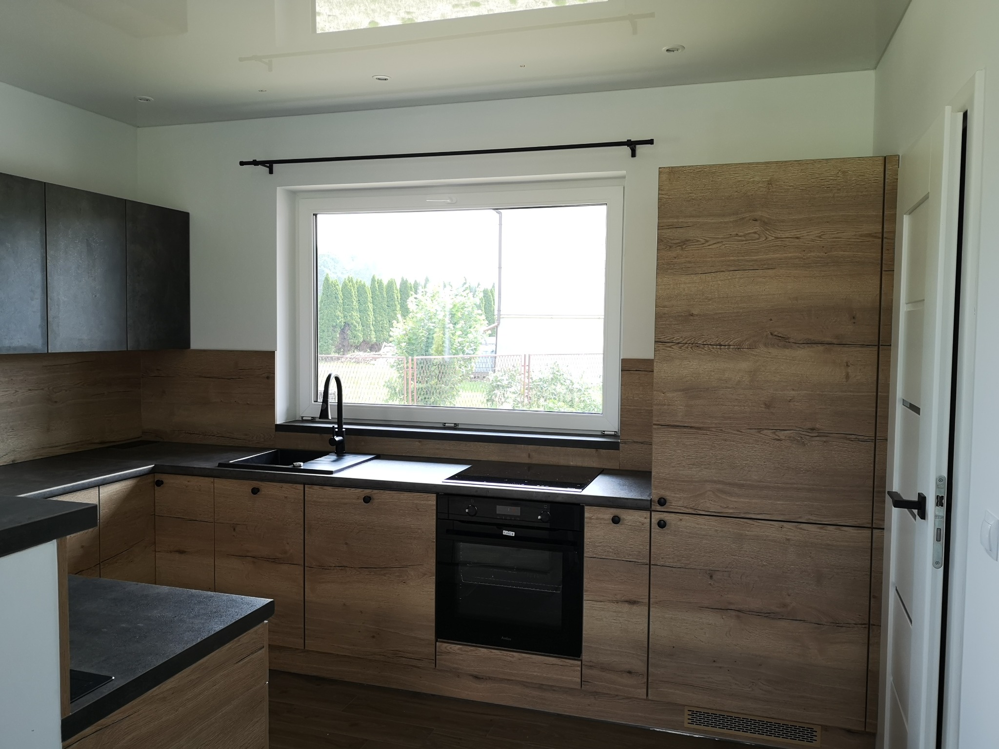 Nowoczesna kuchnia z drewnianymi frontami szafek, czarnym blatem i armaturą, widok na ogród przez okno.