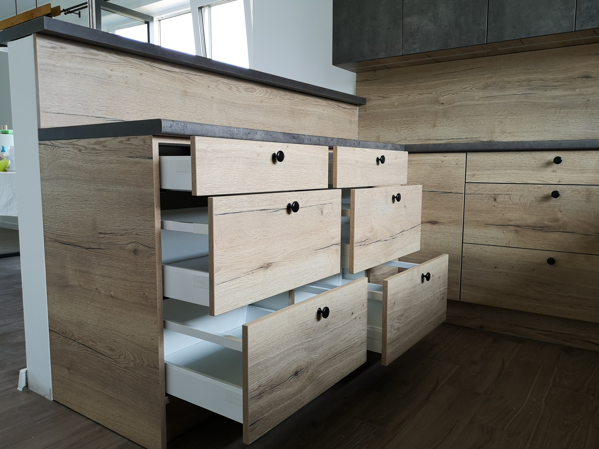Nowoczesna zabudowa kuchenna z drewnopodobnymi frontami i czarnymi uchwytami, szuflady z białym wnętrzem częściowo wysunięte, widoczny blat w kolorze ciemnego betonu.