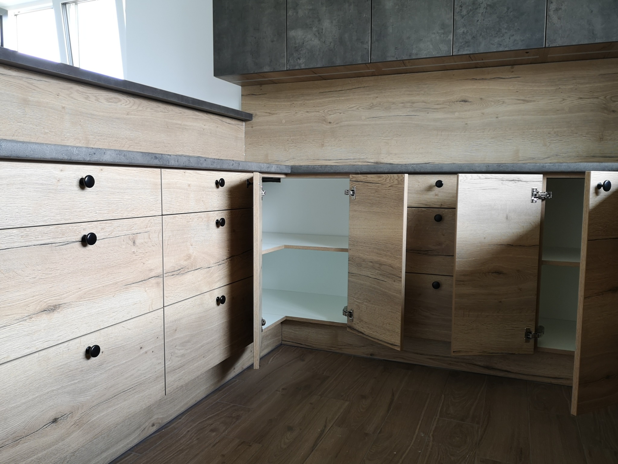 Jasna drewniana zabudowa kuchenna z szarym blatem, czarnymi okrągłymi uchwytami, otwarte szafki narożne z białym wnętrzem.