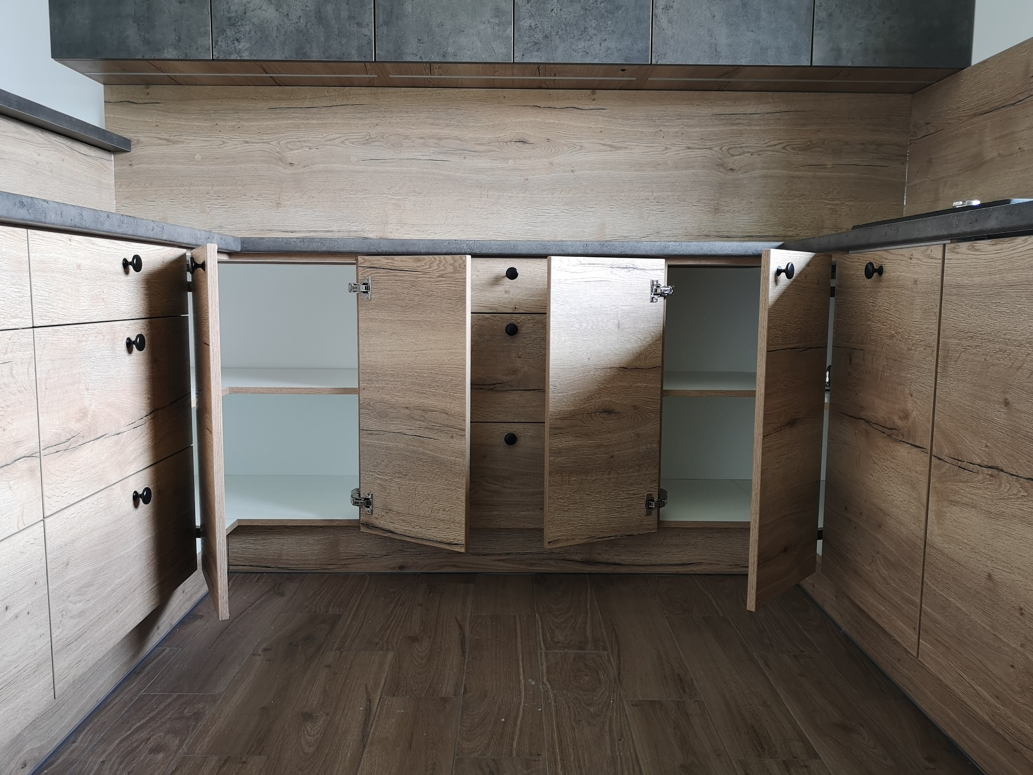 Nowoczesna kuchnia na wymiar z drewnianymi frontami, otwarte szafki ukazują białe wnętrze, ciemny blat i szare płytki nad blatem.