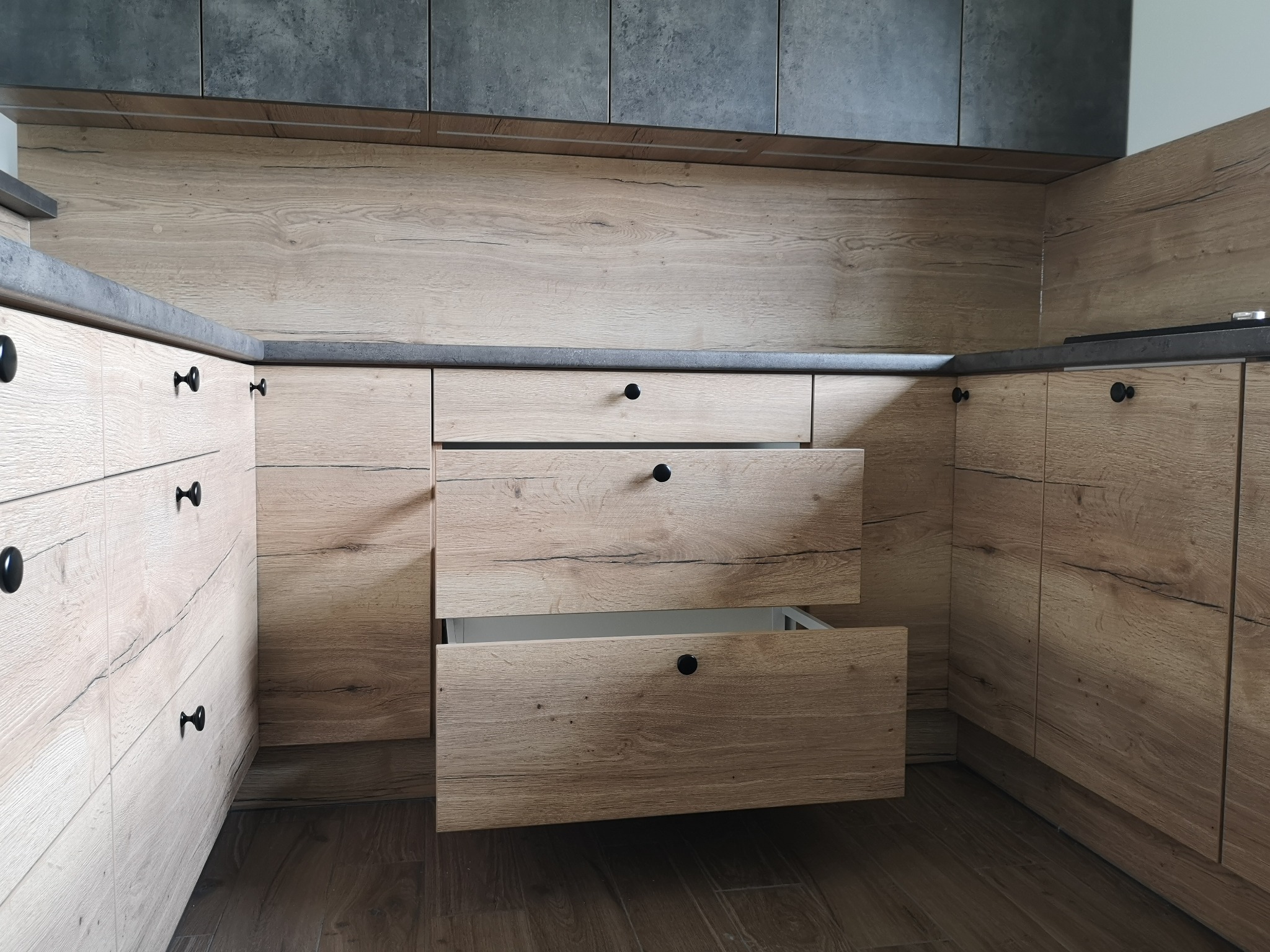 Fragment zabudowy kuchennej z drewnopodobnymi frontami, szarym blatem i otwartą szufladą, eksponującą białe wnętrze.