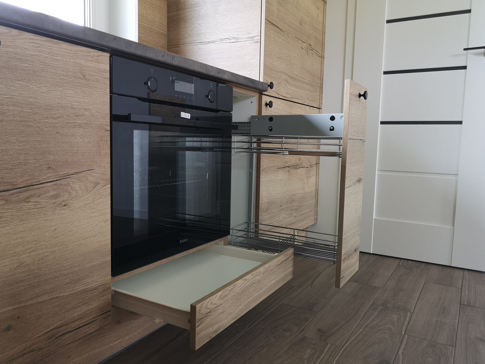 Jasna drewniana zabudowa kuchenna z piekarnikiem i wysuwaną metalową półką cargo, widoczne detale wykończenia i brązowa podłoga.