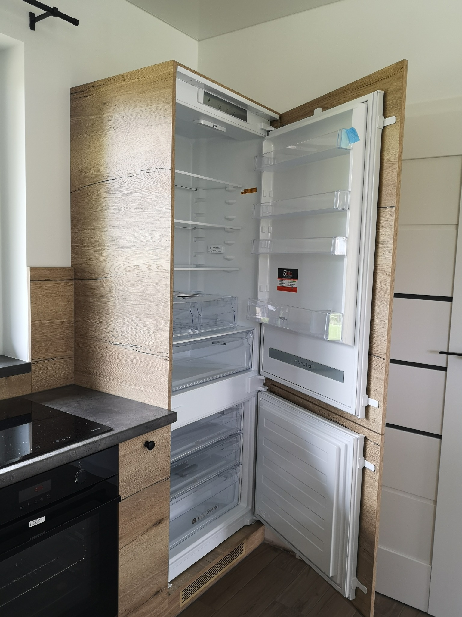 Wnętrze kuchni z zabudowaną lodówką z frontami imitującymi drewno, otwarte drzwi ukazują puste półki i szuflady.