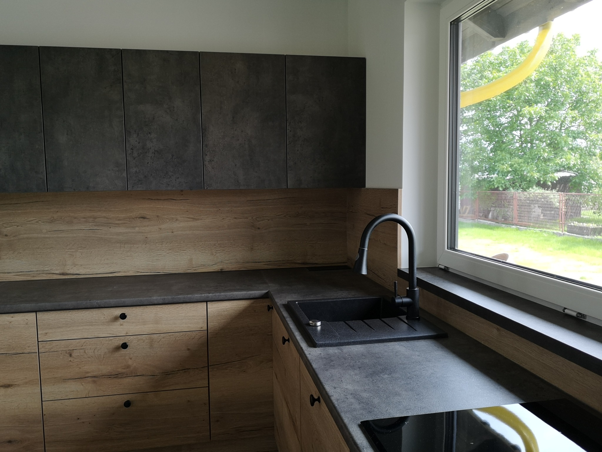 Nowoczesna kuchnia z drewnianymi frontami szafek dolnych i szarymi szafkami górnymi, czarny zlew i bateria, widok na ogród przez okno.