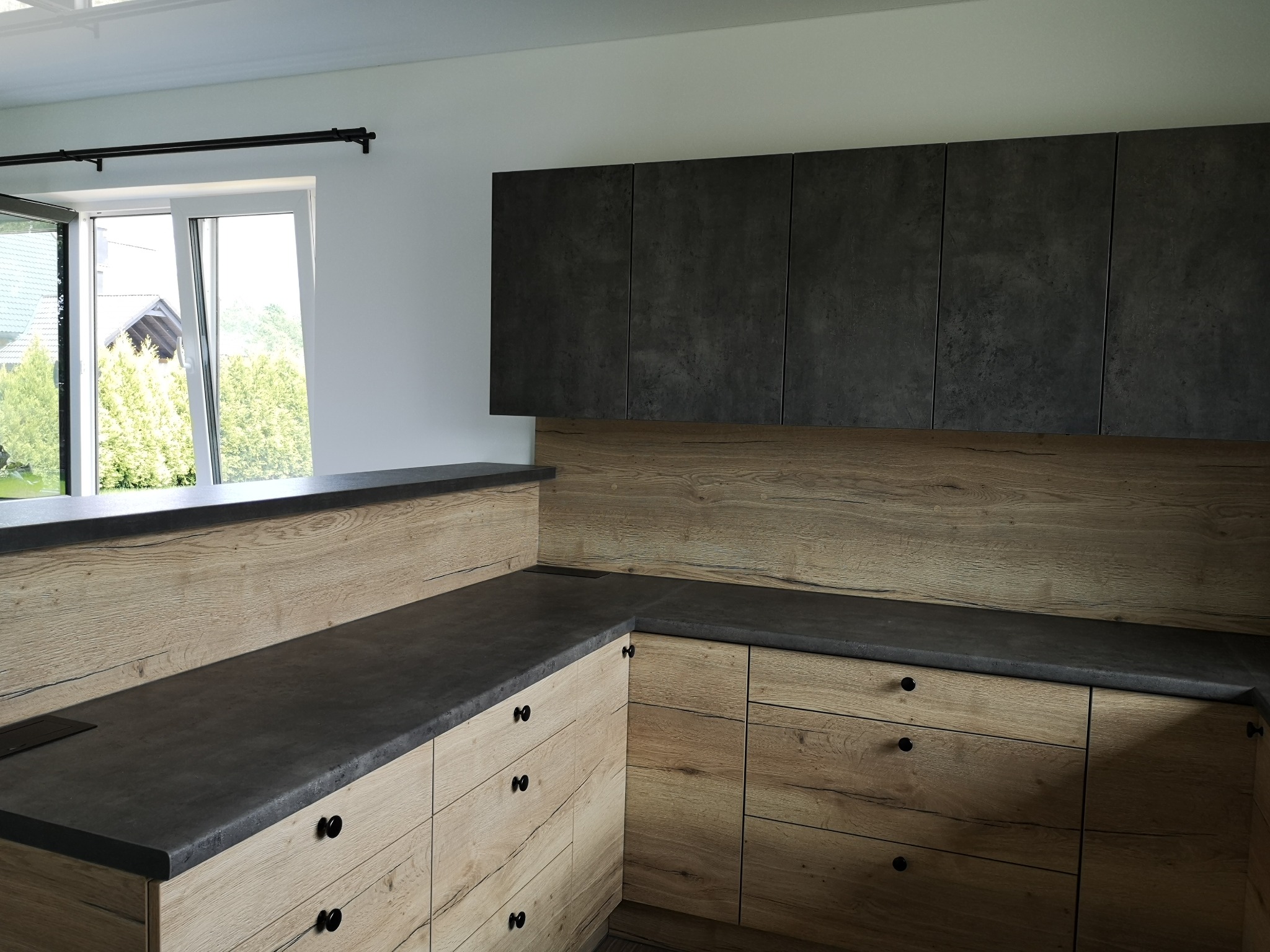Nowoczesna kuchnia z drewnianymi frontami szafek dolnych i ciemnymi szafkami górnymi, czarny blat, widok na ogród przez okno