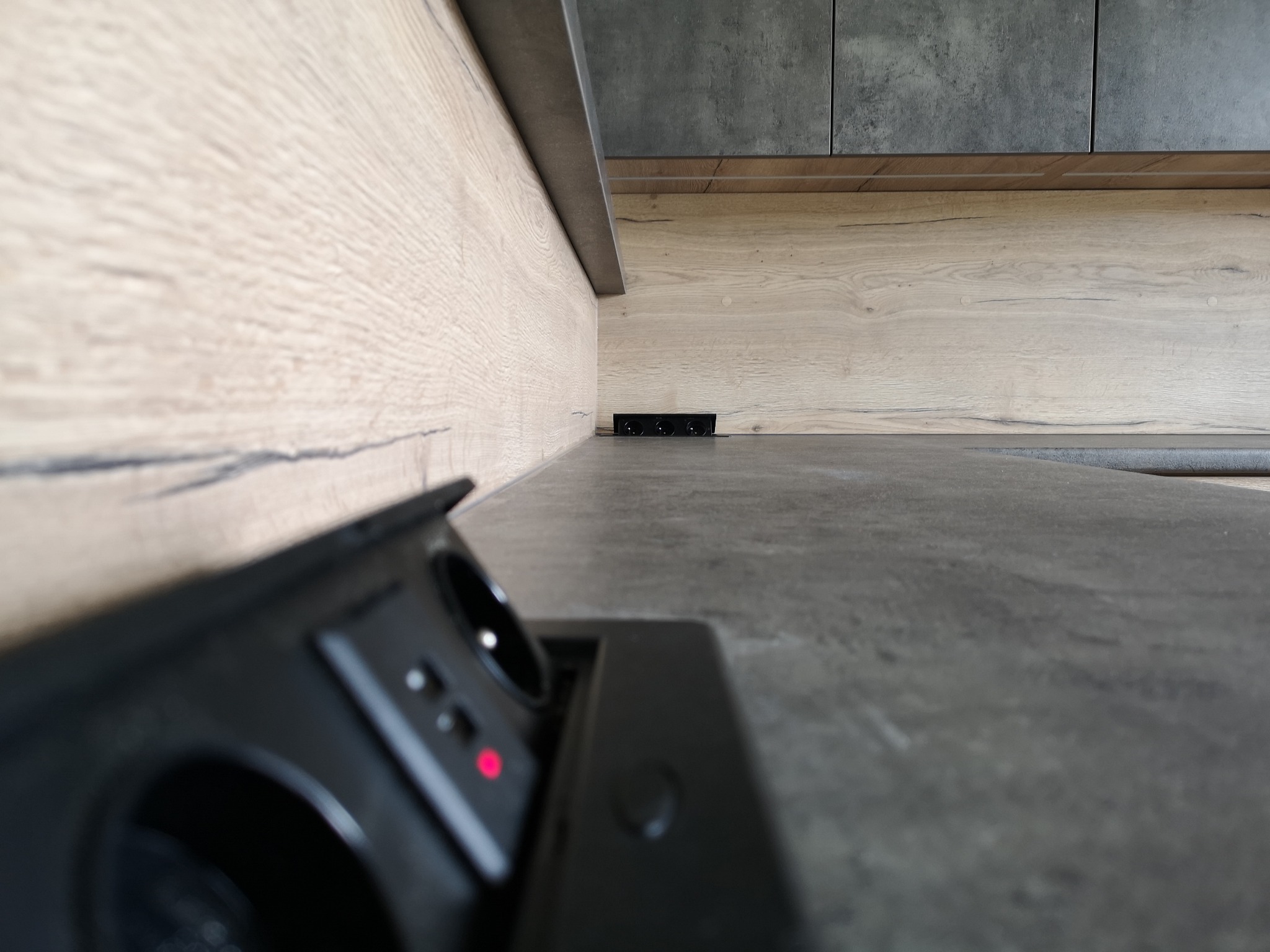 Szary blat kuchenny z ukrytymi gniazdkami, widoczne drewniane panele ścienne i górne szafki w odcieniach szarości i brązu.