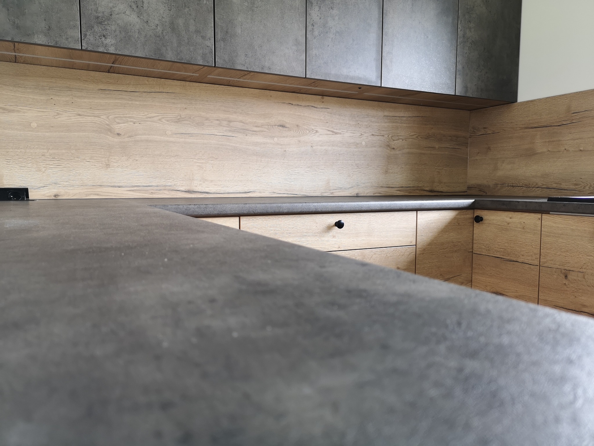 Nowoczesna kuchnia z szarymi szafkami górnymi i drewnopodobnymi frontami dolnymi, widok z bliska na blat z szarego kamienia.