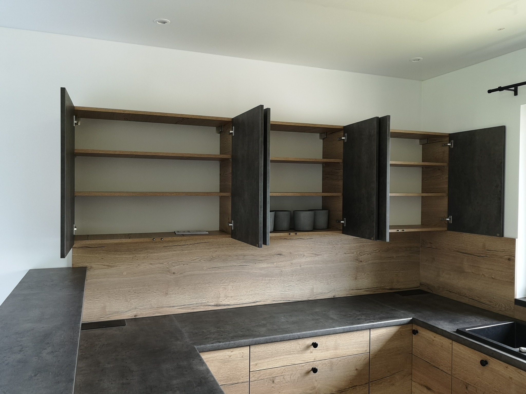 Nowoczesna kuchnia z drewnianymi frontami szafek, szarym blatem i otwartymi szafkami górnymi, w których widać kilka szarych doniczek.