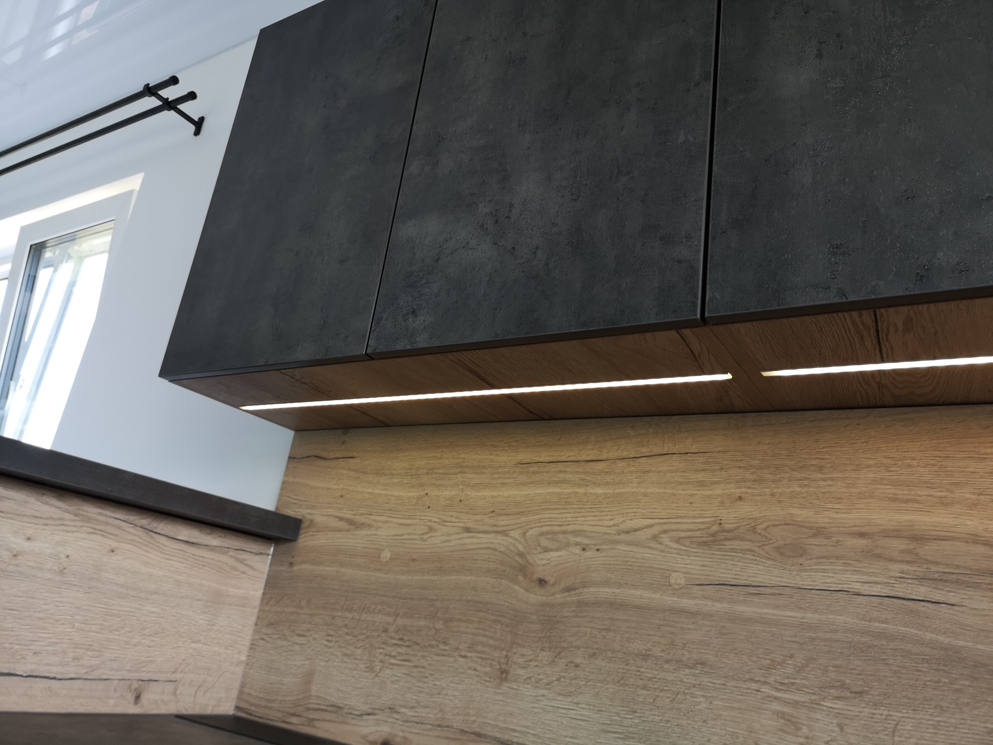 Częściowy widok nowoczesnej zabudowy kuchennej z ciemnymi, matowymi szafkami wiszącymi i drewnopodobnym panelem ściennym z podświetleniem LED.