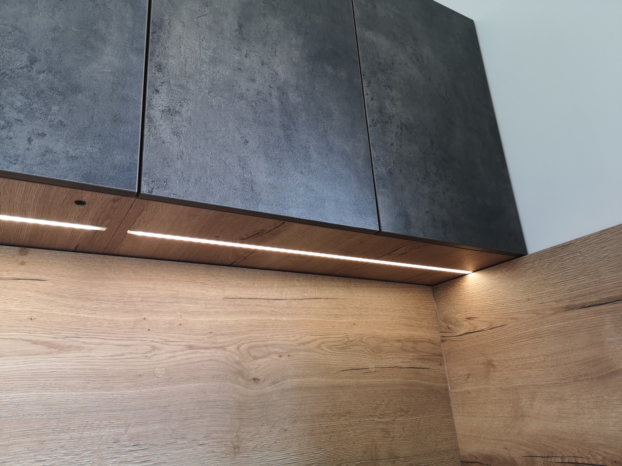 Fragment nowoczesnej zabudowy kuchennej z górnymi szafkami w kolorze grafitowym o matowej fakturze i drewnianymi panelami ściennymi z oświetleniem LED podszafkowym.