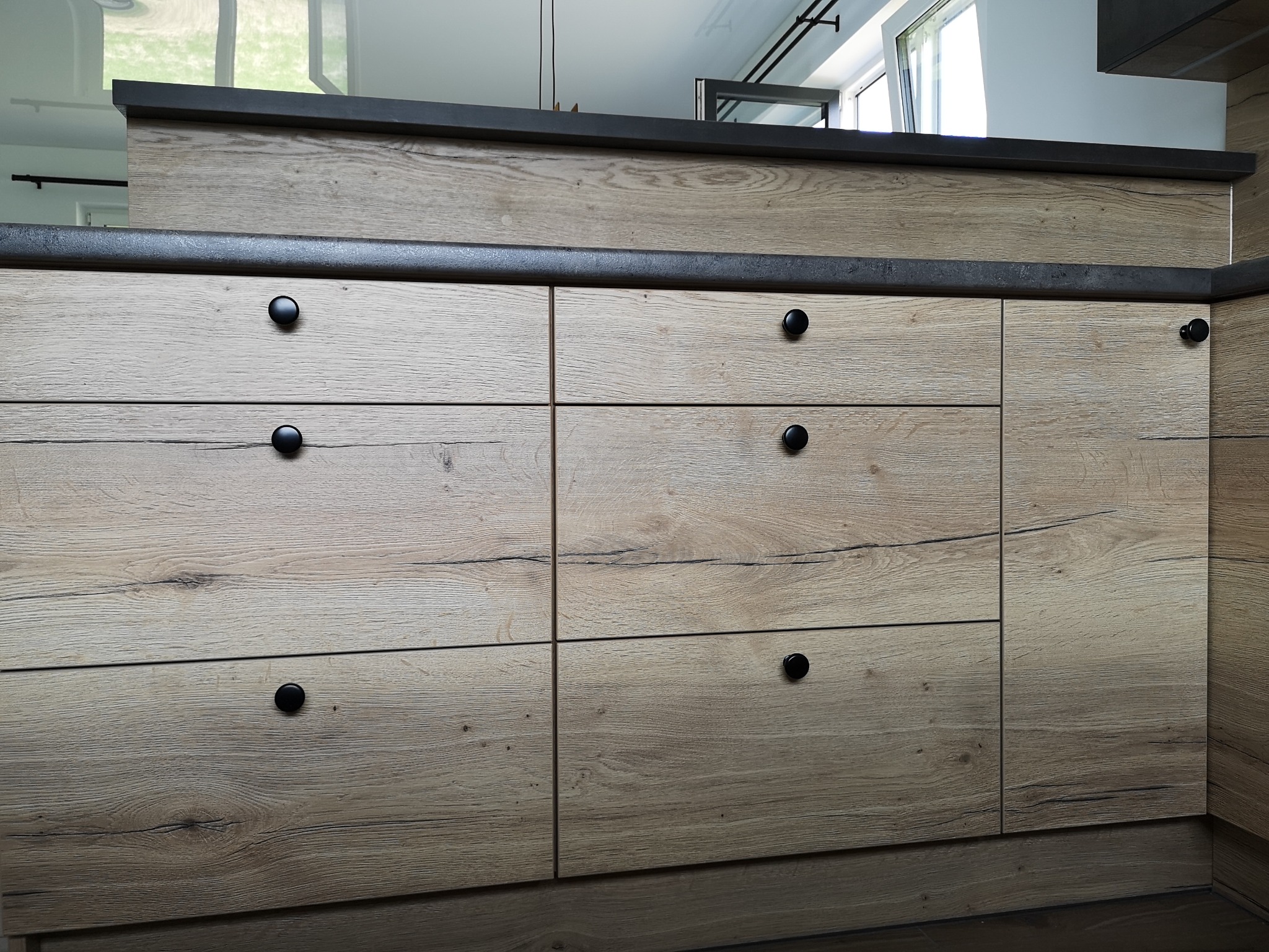 Fronty szafek kuchennych z jasnego drewna z czarnymi, okrągłymi uchwytami, widoczne trzy rzędy szuflad i szafek, czarny blat.