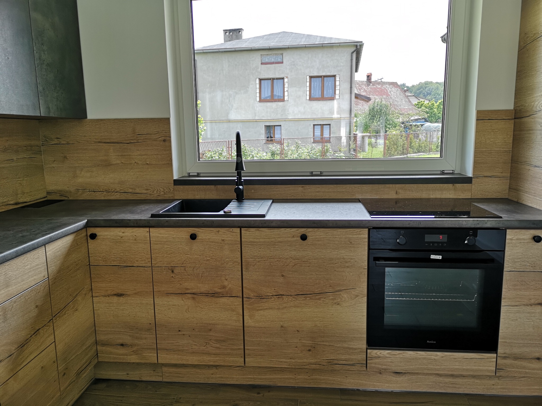 Nowoczesna kuchnia z drewnianymi frontami szafek, czarnym zlewem i baterią, piekarnikiem Amica oraz widokiem na budynek mieszkalny za oknem.