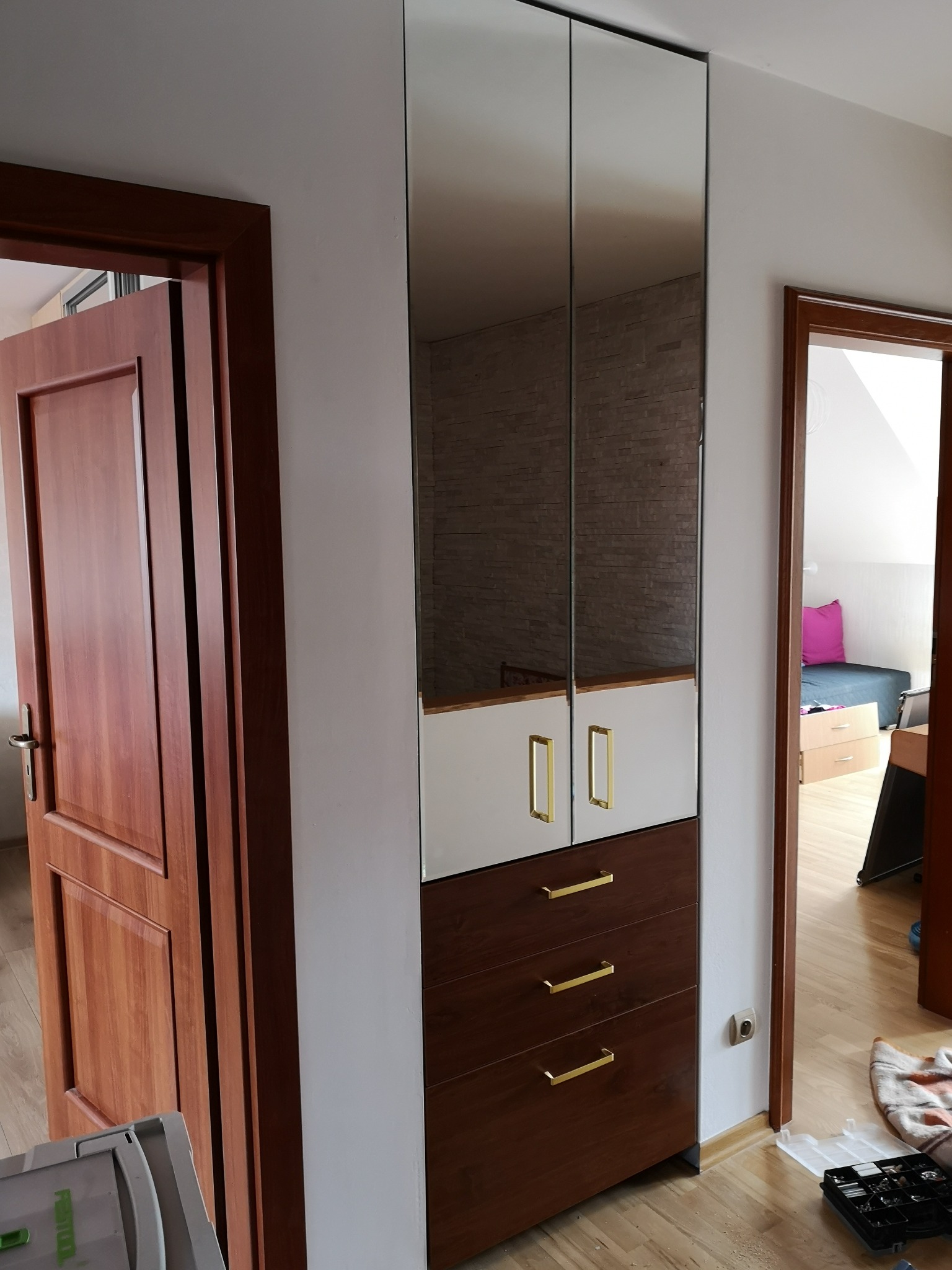 Wysoka szafa z lustrzanymi drzwiami, białymi i brązowymi frontami szuflad, ze złotymi uchwytami, widziana w korytarzu obok otwartych drzwi do pokoju.