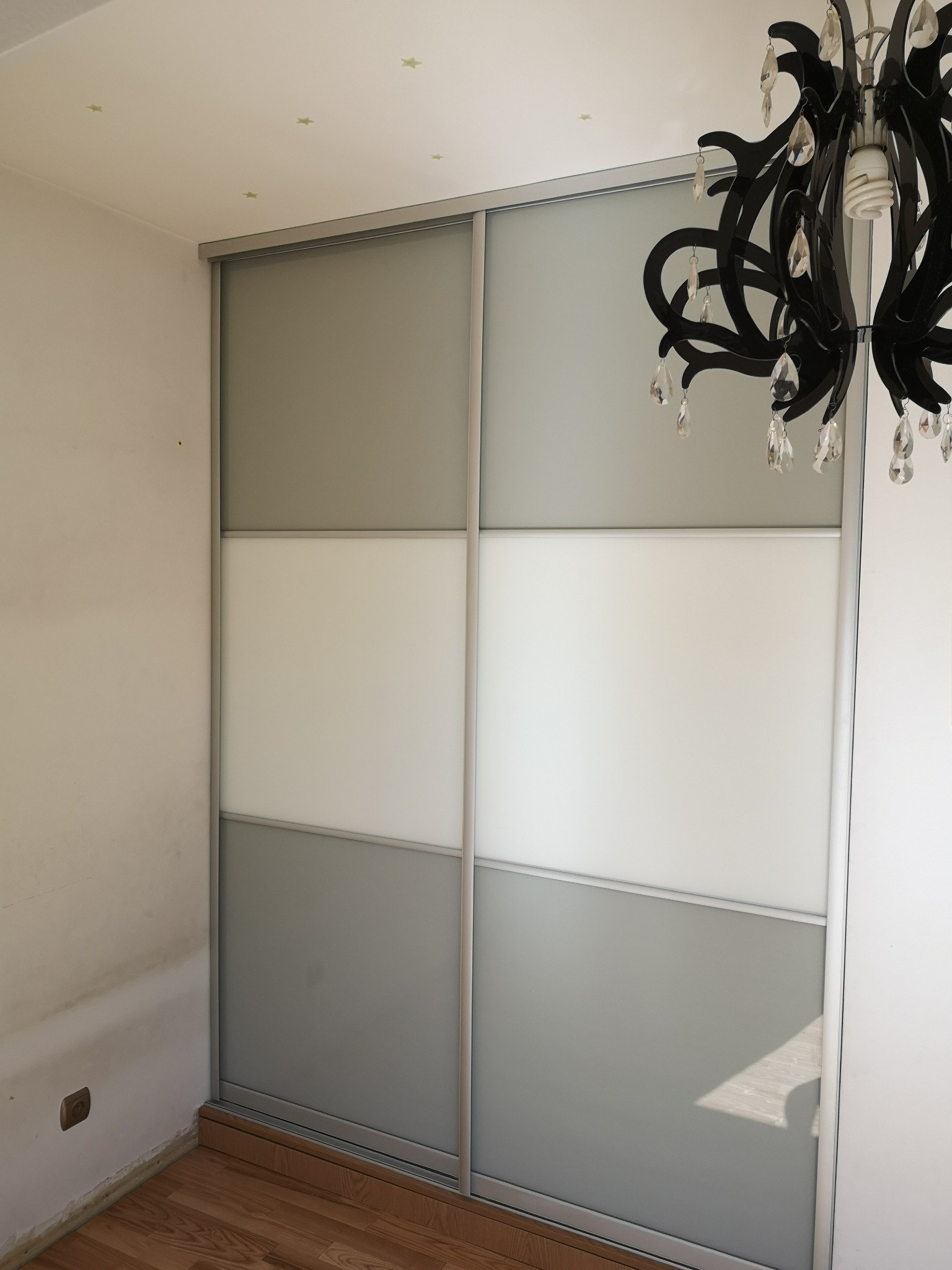 Czteropanelowa szafa przesuwna z szarymi i białymi panelami, oświetlona przez czarny żyrandol z kryształkami, w pokoju z drewnianą podłogą i dekoracyjnymi gwiazdkami na suficie.