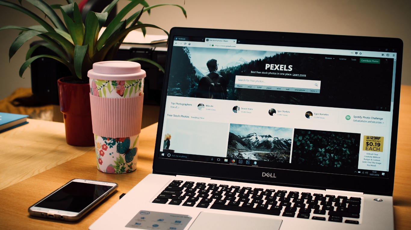 Laptop Dell z otwartą stroną Pexels, obok kubek termiczny w kwiaty i telefon na biurku z drewnianym blatem, w tle roślina doniczkowa.