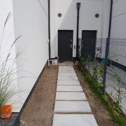 Finex-Bud - Chodnik z betonowych płyt prowadzący do drzwi budynku, widoczne świeżo posadzone trawy i siatka ogrodzeniowa.