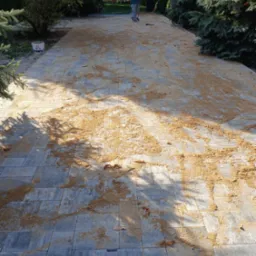 Świeżo ułożona szara kostka brukowa z piaskiem do wypełniania fug, widoczny pracownik w oddali i drzewa w tle.