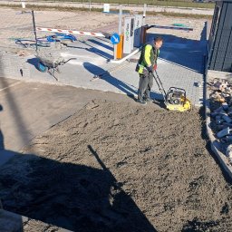 Finex-Bud - Pracownik w kamizelce odblaskowej zagęszcza piasek ubijarką wibracyjną przed ułożeniem kostki brukowej na parkingu; w tle szlaban, wózek z kostką i budynki.