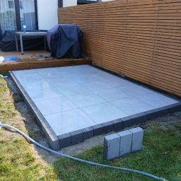 Finex-Bud - Układanie tarasu z szarych płytek, obrzeża z ciemnej kostki, widoczny fragment drewnianego ogrodzenia i zraszacz na trawie.