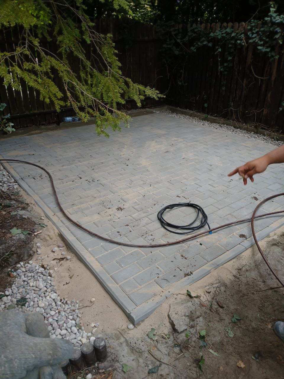 Ułożona szara kostka brukowa na piaskowym podłożu, widoczny kabel i fragment dłoni wskazującej na nawierzchnię, tło stanowi drewniany płot i zieleń drzew.