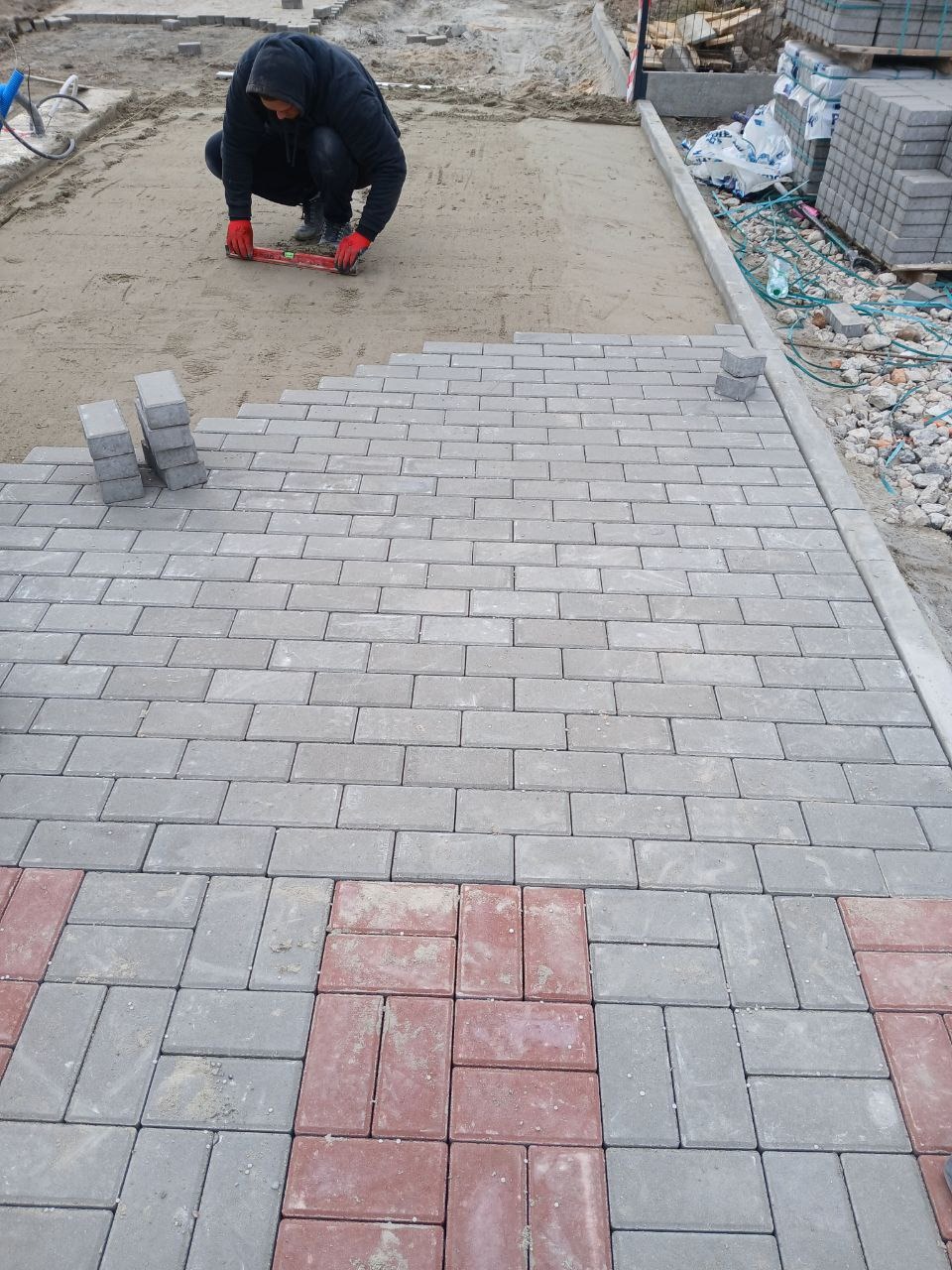 Pracownik w czerwonych rękawicach klęczy, poziomując układaną szarą kostkę brukową z wstawkami z czerwonej kostki. W tle stos kostki na palecie i przygotowane podłoże.