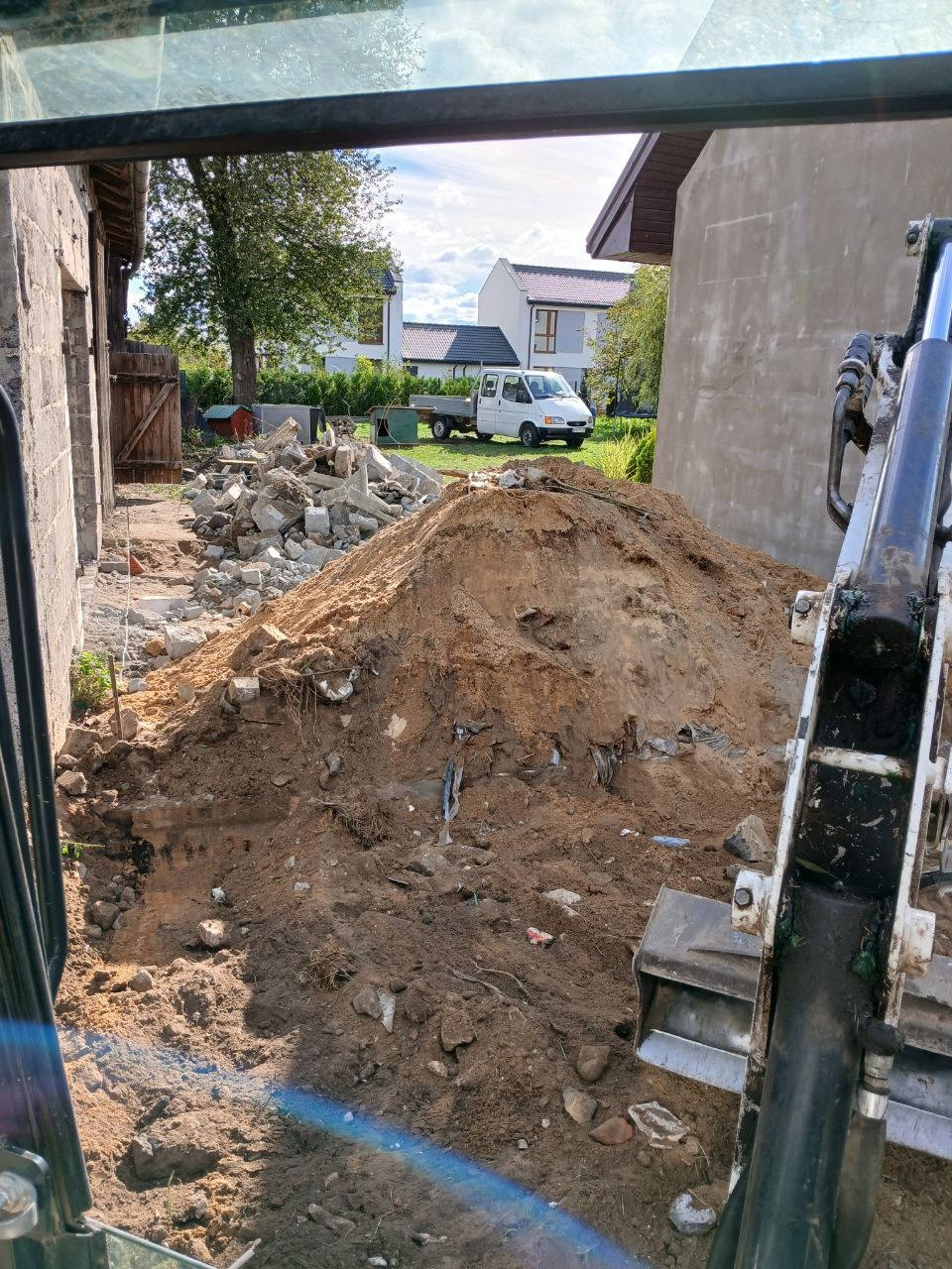 Kopuła ziemi z gruzem obok budynku, widok z kabiny koparki, w tle zaparkowany biały samochód dostawczy.