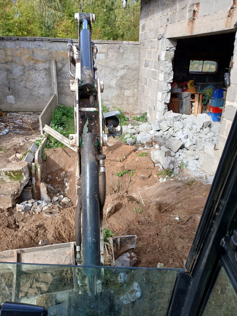 Widok z kabiny koparki na rozbierany budynek z gruzem i wykopem w ziemi.