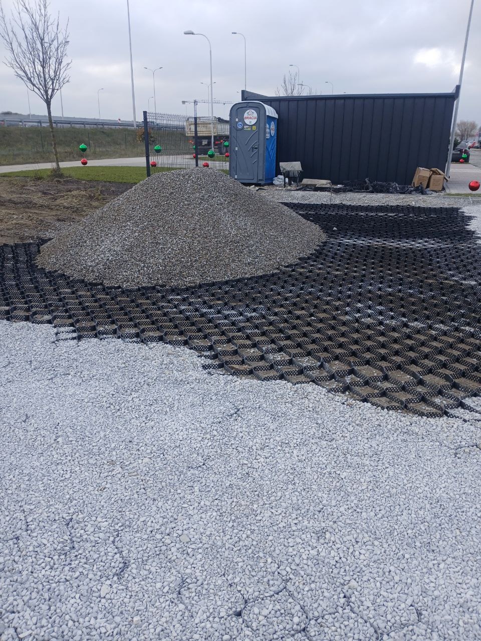 Przygotowanie podłoża: czarne plastikowe kratki częściowo wypełnione szarym żwirem, obok pryzma żwiru, w tle przenośna toaleta i ogrodzenie na pochmurny dzień.