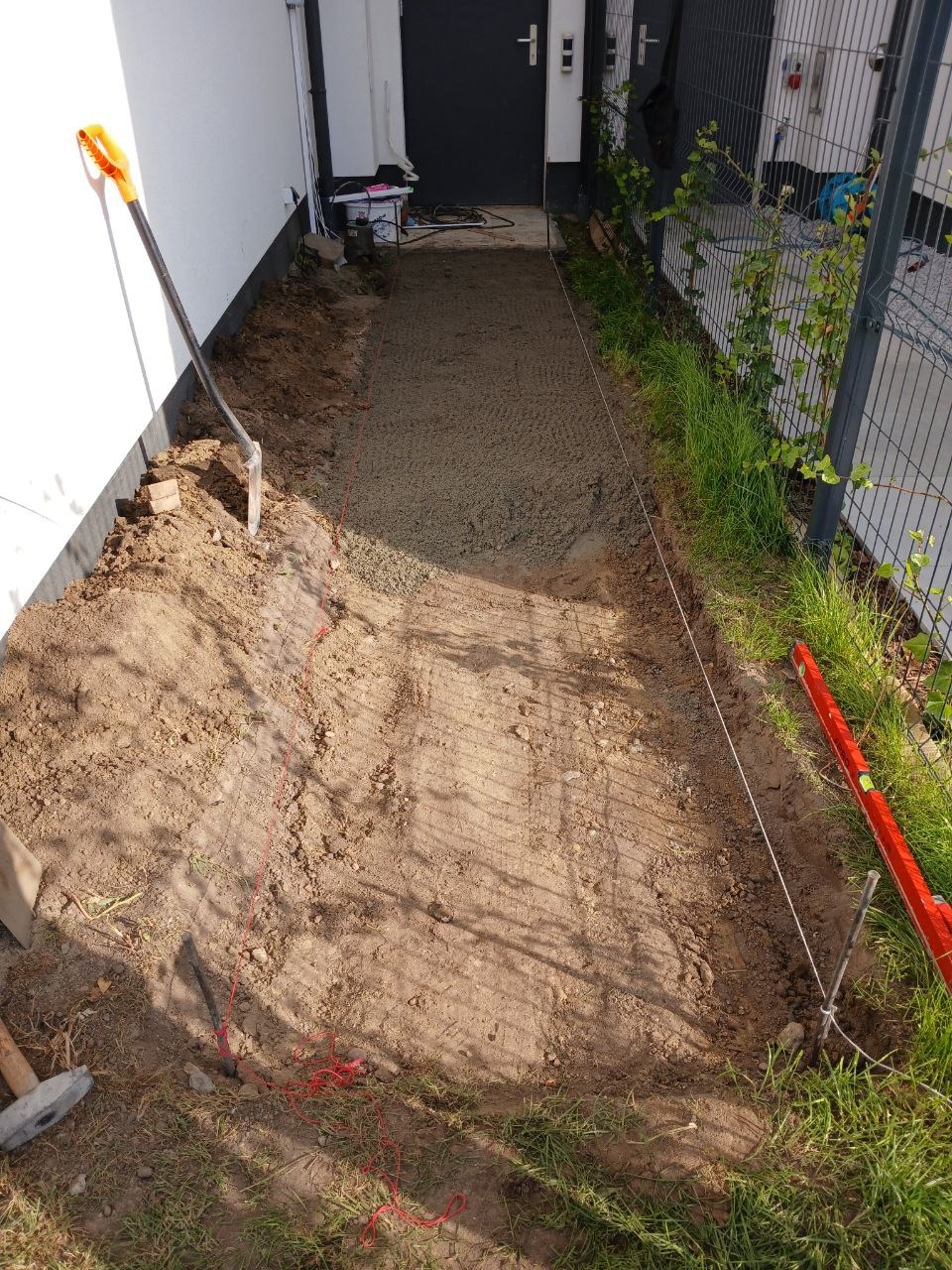 Przygotowanie podłoża pod ścieżkę: wyrównany teren z podsypką, wytyczone sznurki, łopata oparta o ścianę budynku, poziomica przy ogrodzeniu.