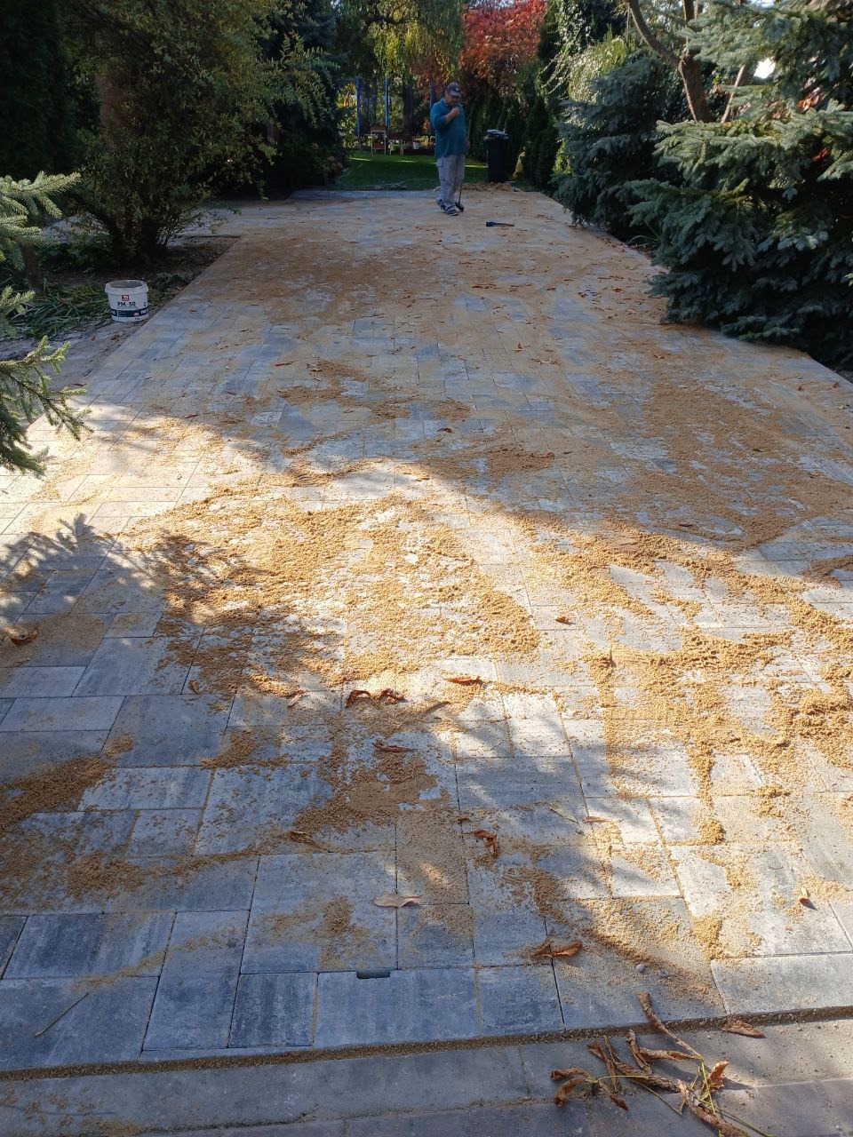 Świeżo ułożona szara kostka brukowa z piaskiem do wypełniania fug, widoczny pracownik w oddali i drzewa w tle.