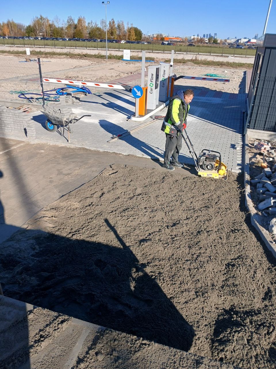 Pracownik w kamizelce odblaskowej zagęszcza piasek ubijarką wibracyjną przed ułożeniem kostki brukowej na parkingu; w tle szlaban, wózek z kostką i budynki.