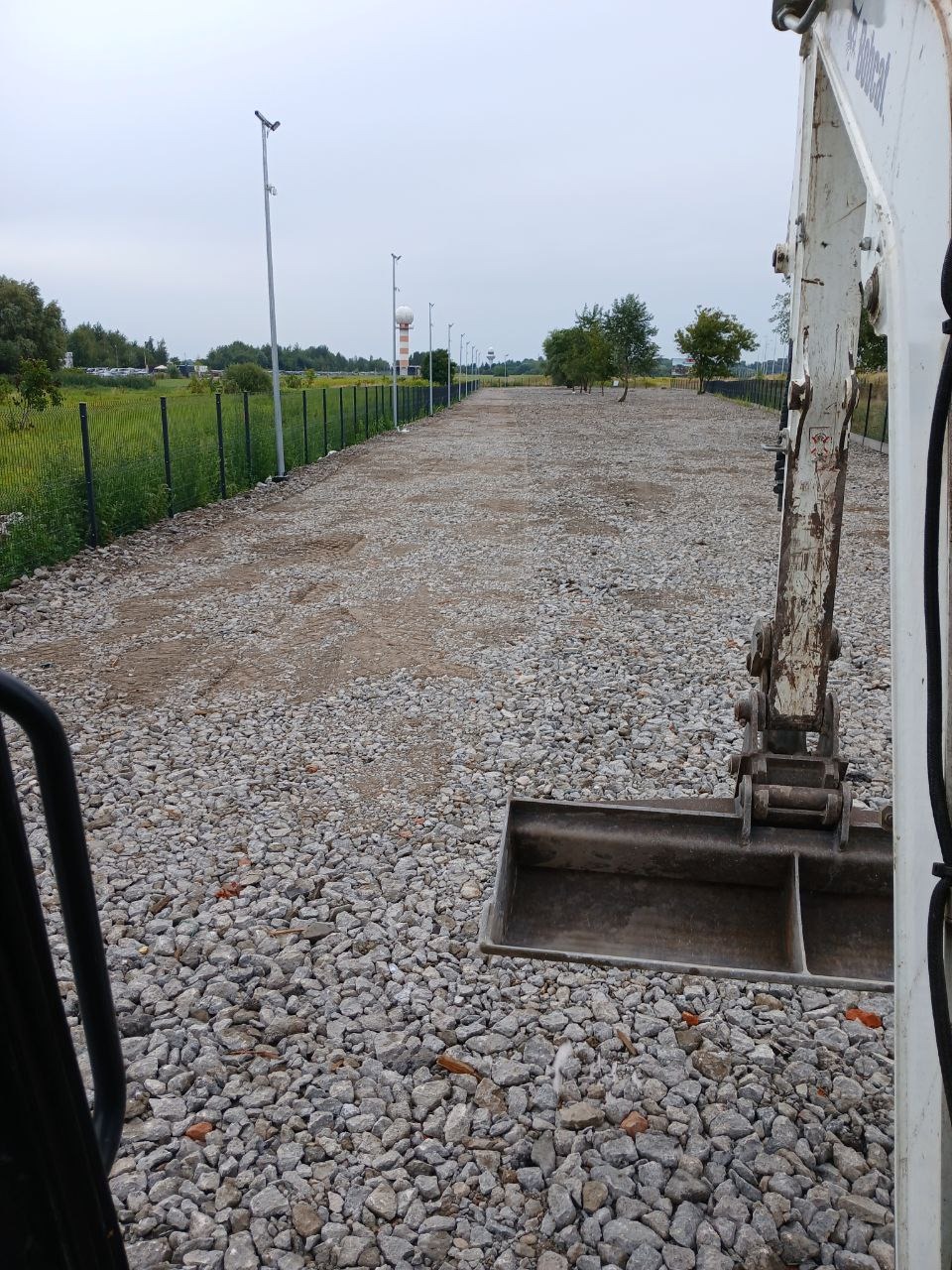 Przygotowanie terenu pod budowę: mini koparka z łyżką na żwirowym podłożu, widoczny fragment ogrodzenia z siatki i słupy oświetleniowe w tle.