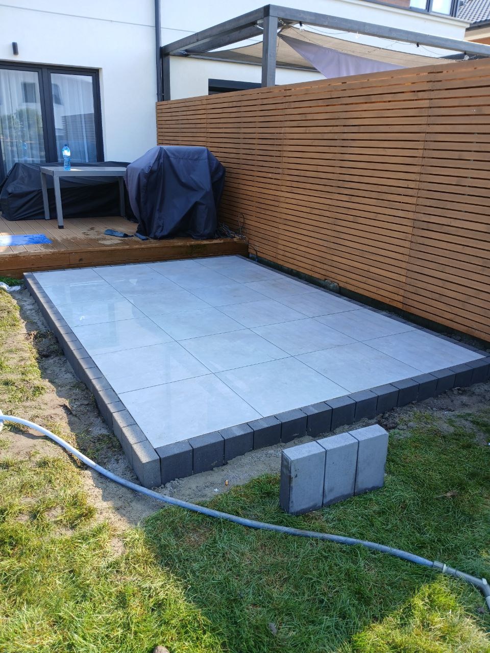 Układanie tarasu z szarych płytek, obrzeża z ciemnej kostki, widoczny fragment drewnianego ogrodzenia i zraszacz na trawie.