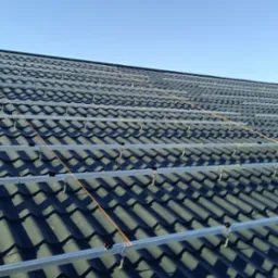 Montaż szyn pod panele fotowoltaiczne na dachu pokrytym ciemną dachówką ceramiczną, widoczne mocowania i linki poziomujące, błękitne niebo w tle.