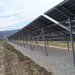 BRIGHT ECO ENERGY SPÓŁKA Z OGRANICZONĄ ODPOWIEDZIALNOŚCIĄ - Rząd paneli słonecznych zamontowanych na metalowej konstrukcji, widok z dołu, z podsypką z jasnego żwiru i błotnistym terenem obok, w tle zabudowania i wzgórza, pochmurne niebo.