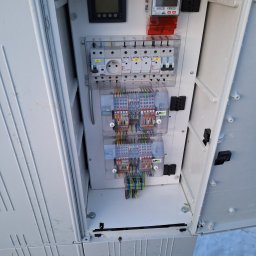 BRIGHT ECO ENERGY SPÓŁKA Z OGRANICZONĄ ODPOWIEDZIALNOŚCIĄ - Otwarta biała szafa elektryczna z widocznymi bezpiecznikami, licznikiem, gniazdem oraz kolorowymi przewodami podłączonymi do listew zaciskowych. Na zewnątrz szafy widoczny śnieg.