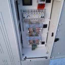 Otwarta biała szafa elektryczna z widocznymi bezpiecznikami, licznikiem, gniazdem oraz kolorowymi przewodami podłączonymi do listew zaciskowych. Na zewnątrz szafy widoczny śnieg.