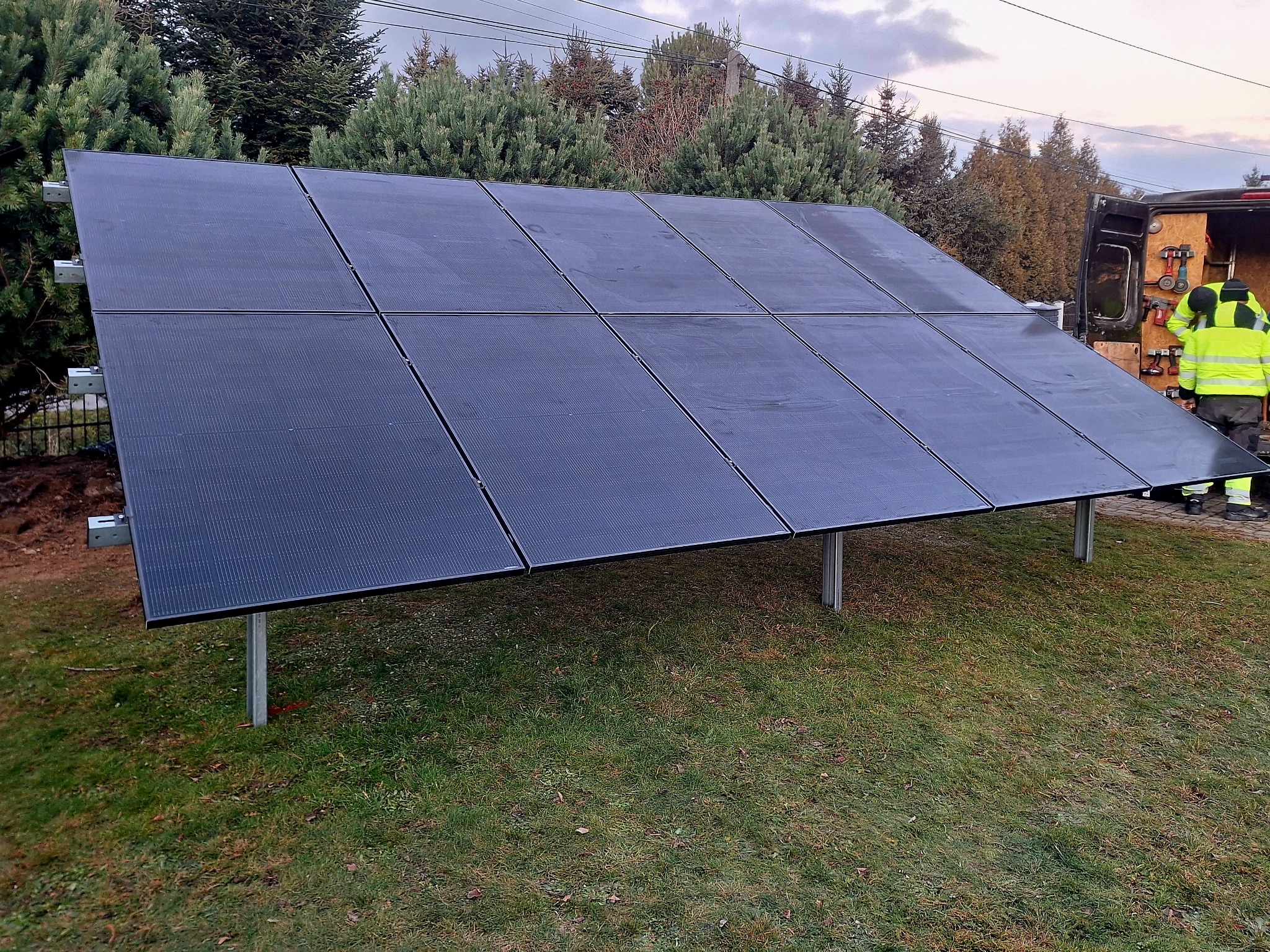Czarny panel słoneczny na metalowej konstrukcji, zamontowany na trawniku, obok zaparkowany samochód z narzędziami, w tle drzewa i pochmurne niebo.