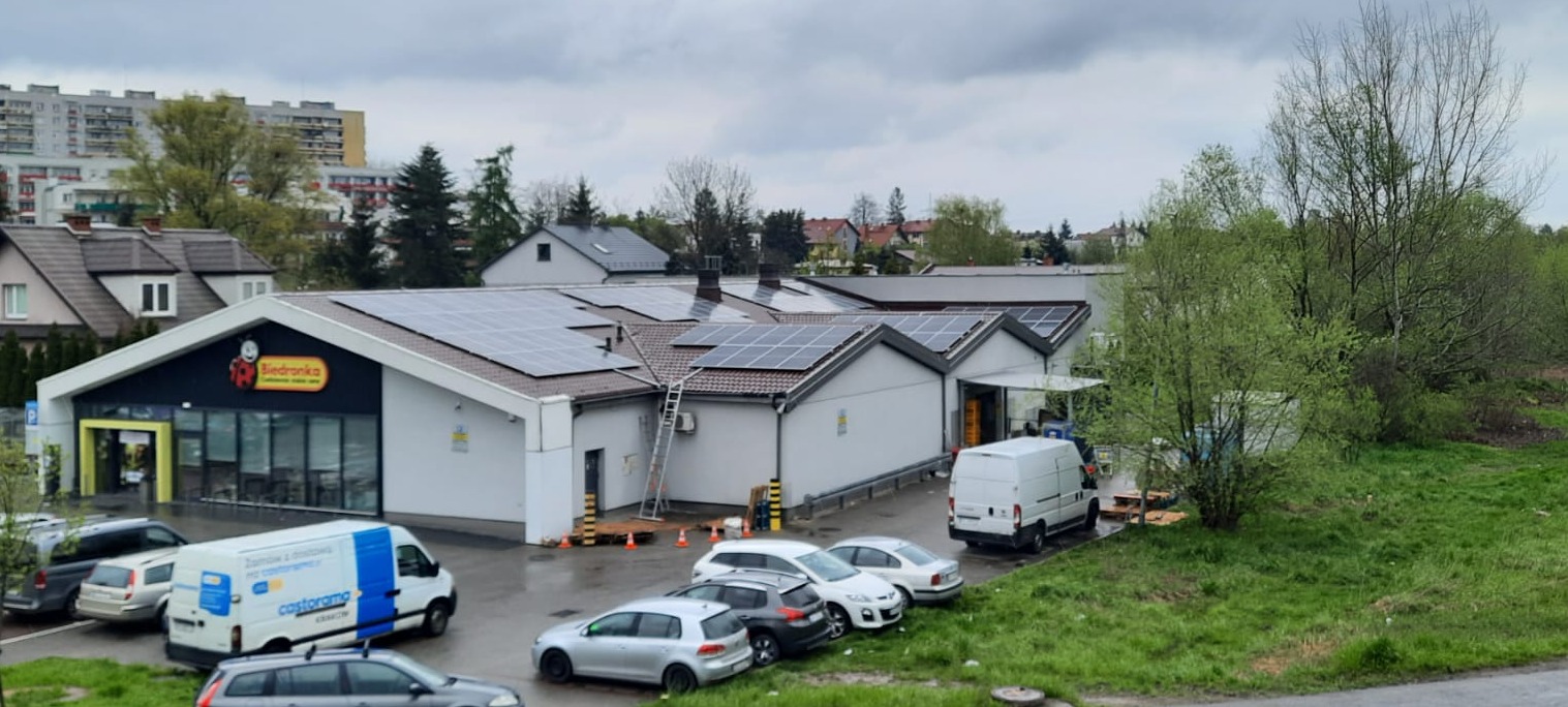 Budynek sklepu Biedronka z zainstalowanymi panelami słonecznymi na dachu, drabina oparta o ścianę, kilka samochodów na parkingu i zieleń w tle.