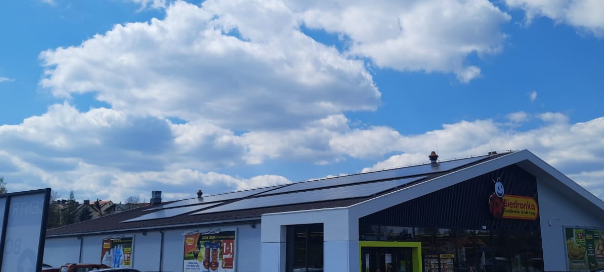 Panele fotowoltaiczne na dachu budynku sklepu Biedronka w słoneczny dzień z chmurami.
