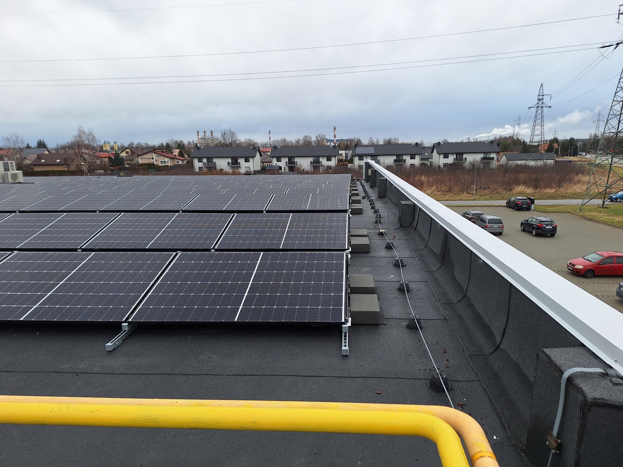 Rząd paneli słonecznych na płaskim dachu budynku komercyjnego, widok z góry, z żółtą rurą zabezpieczającą w pierwszym planie i budynkami mieszkalnymi w tle.