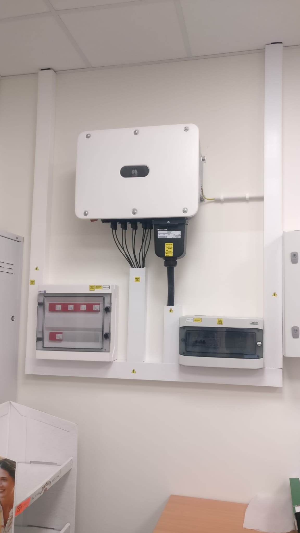 Biały inwerter z czarnymi kablami podłączony do skrzynki elektrycznej na białej ścianie, widoczne ostrzeżenia o napięciu.