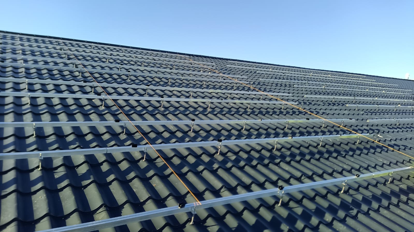 Montaż szyn pod panele fotowoltaiczne na dachu pokrytym ciemną dachówką ceramiczną, widoczne mocowania i linki poziomujące, błękitne niebo w tle.