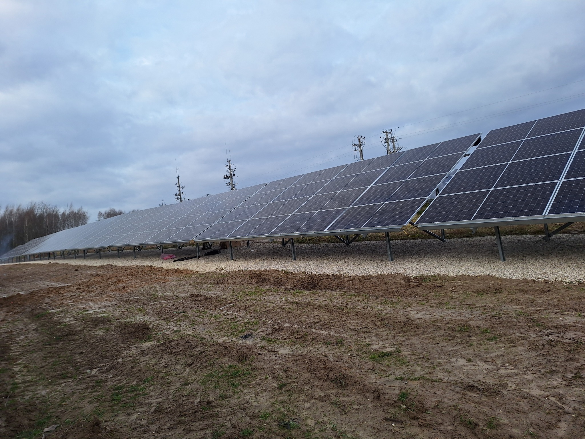 Długa linia paneli słonecznych na metalowych wspornikach ustawionych na żwirowym podłożu, z widocznymi słupami energetycznymi w tle pod pochmurnym niebem.