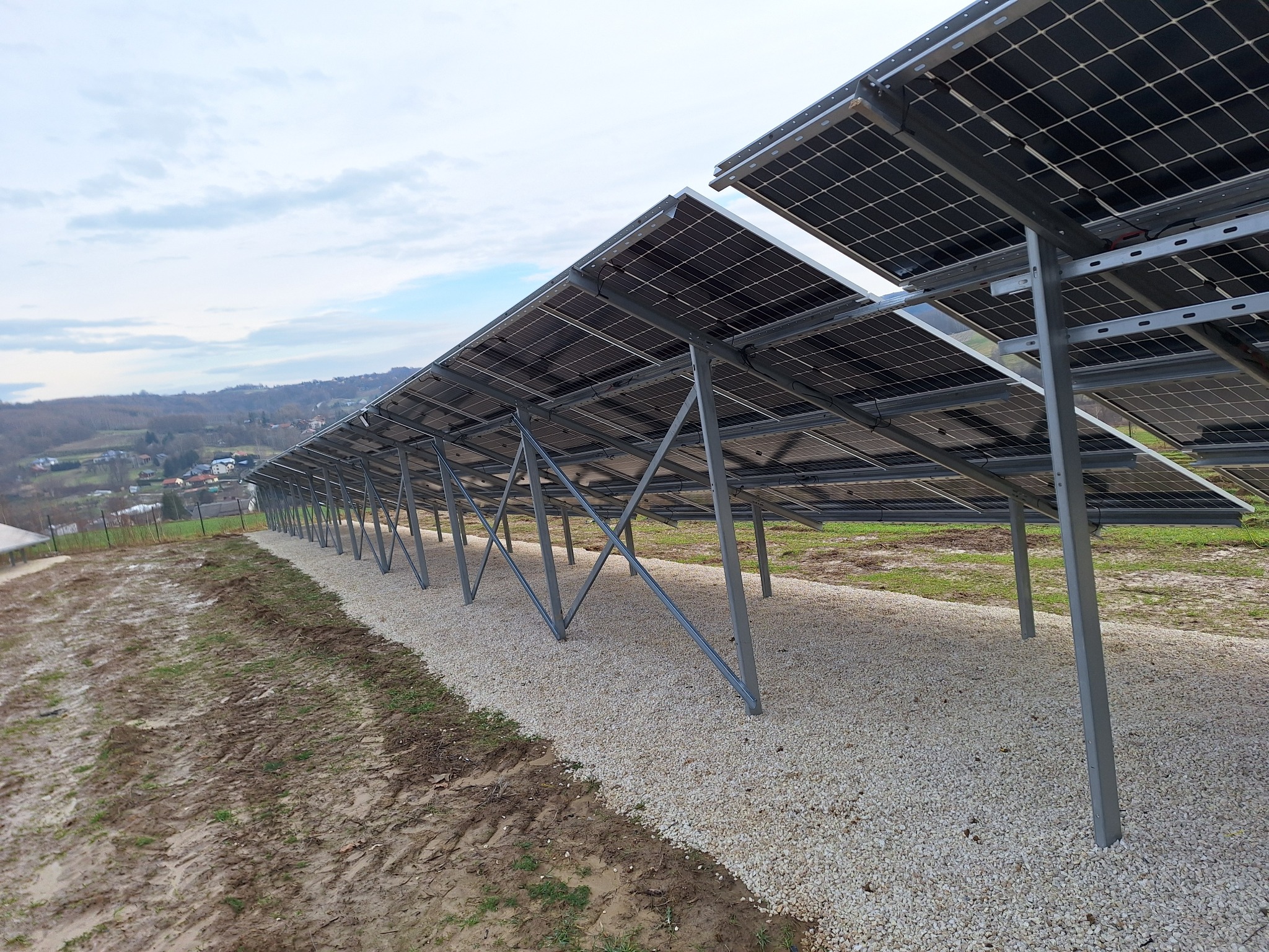 Rząd paneli słonecznych zamontowanych na metalowej konstrukcji, widok z dołu, z podsypką z jasnego żwiru i błotnistym terenem obok, w tle zabudowania i wzgórza, pochmurne niebo.