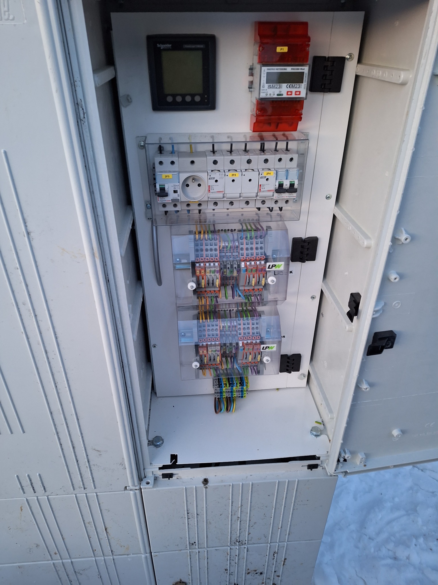 Otwarta biała szafa elektryczna z widocznymi bezpiecznikami, licznikiem, gniazdem oraz kolorowymi przewodami podłączonymi do listew zaciskowych. Na zewnątrz szafy widoczny śnieg.