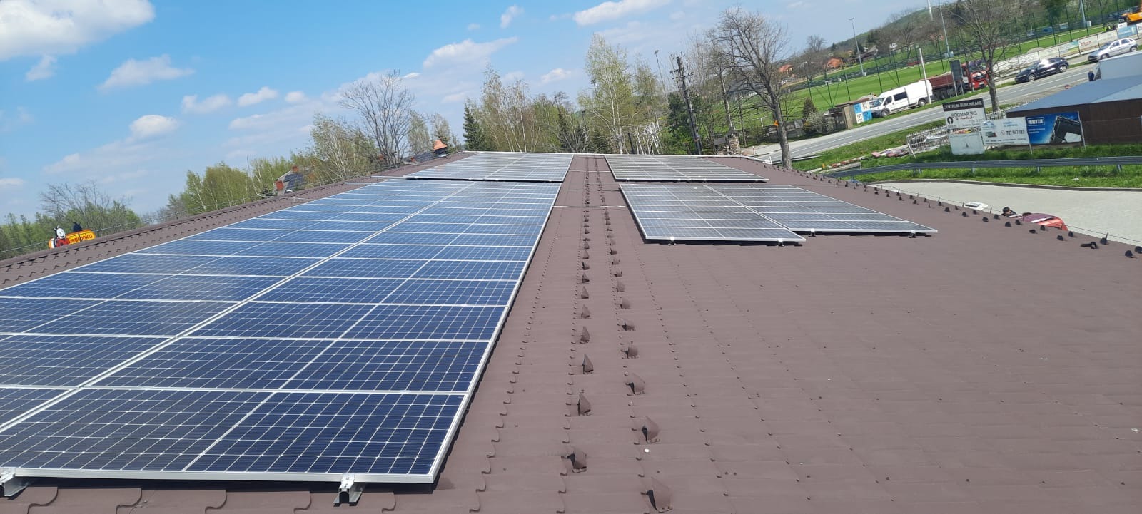 Panele słoneczne na brązowym dachu, widok z góry, błękitne niebo z chmurami w tle, w oddali zabudowania i drzewa.